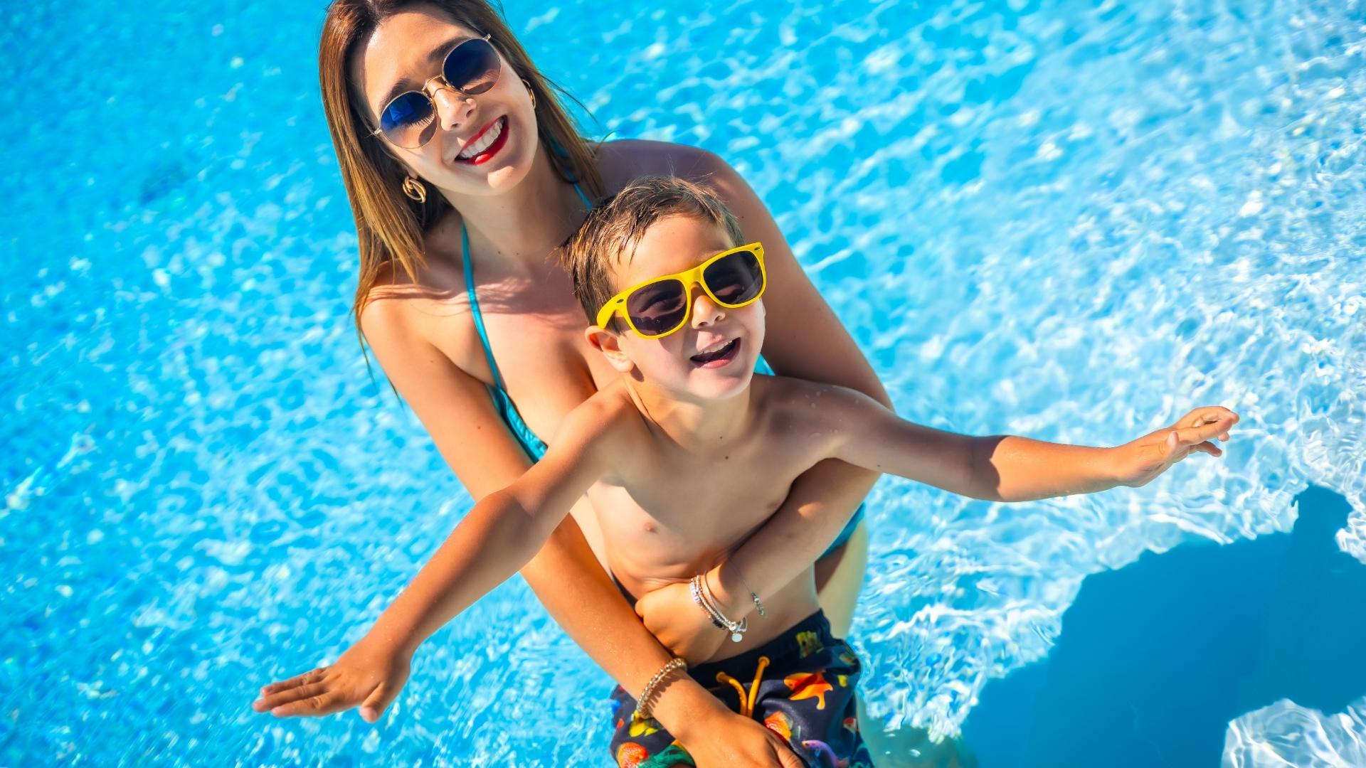 Madre e figlio sorridono felici in piscina, indossando occhiali da sole colorati.