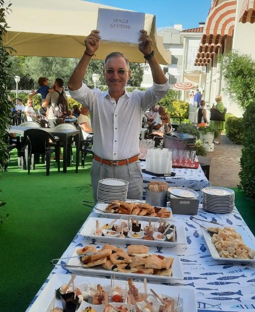 Smiling man holds 'Gluten Free' sign at outdoor buffet, lively and welcoming atmosphere.