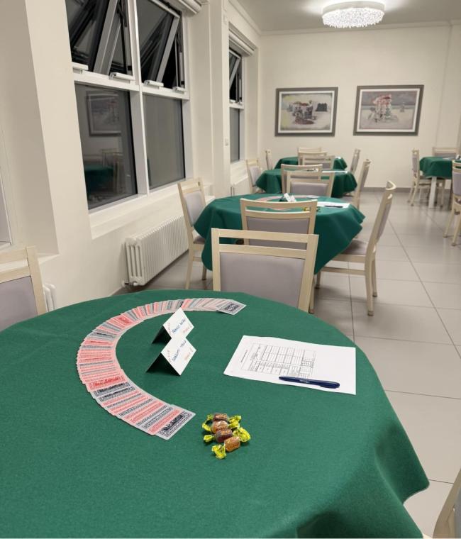 Sala accogliente, tavoli verdi con carte da gioco, caramelle e atmosfera di competizione amichevole.