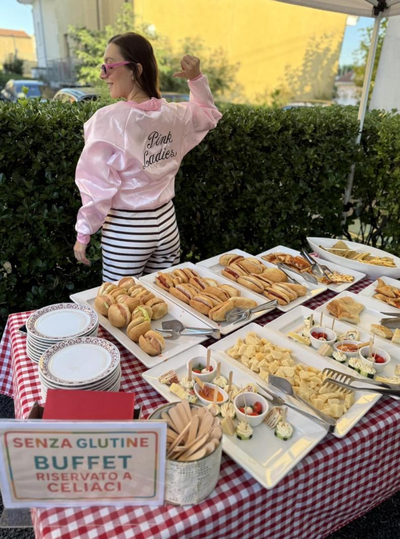 Buffet senza glutine all'aperto, atmosfera anni '50, giacca rosa 'Pink Ladies', energia vivace.
